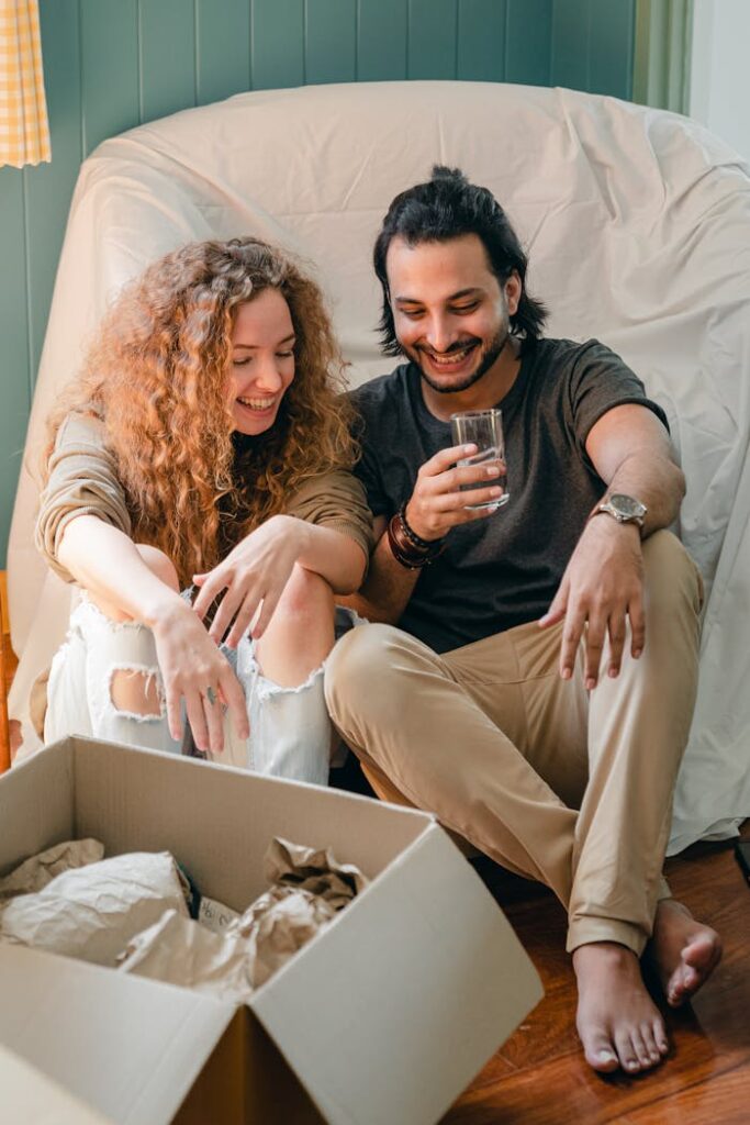 Happy ethnic barefoot man sitting with glass of water on comfortable sofa while talking to positive girlfriend near open cardboard box with parchment at home